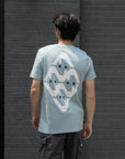 Person wearing a light blue t-shirt with a geometric design, standing against a dark brick wall.