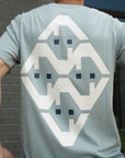 Person wearing a light blue t-shirt with a geometric pattern on the back, standing against a brick wall.