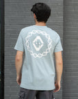 Person wearing a light blue t-shirt with a white graphic design on the back, standing against a gray brick wall.