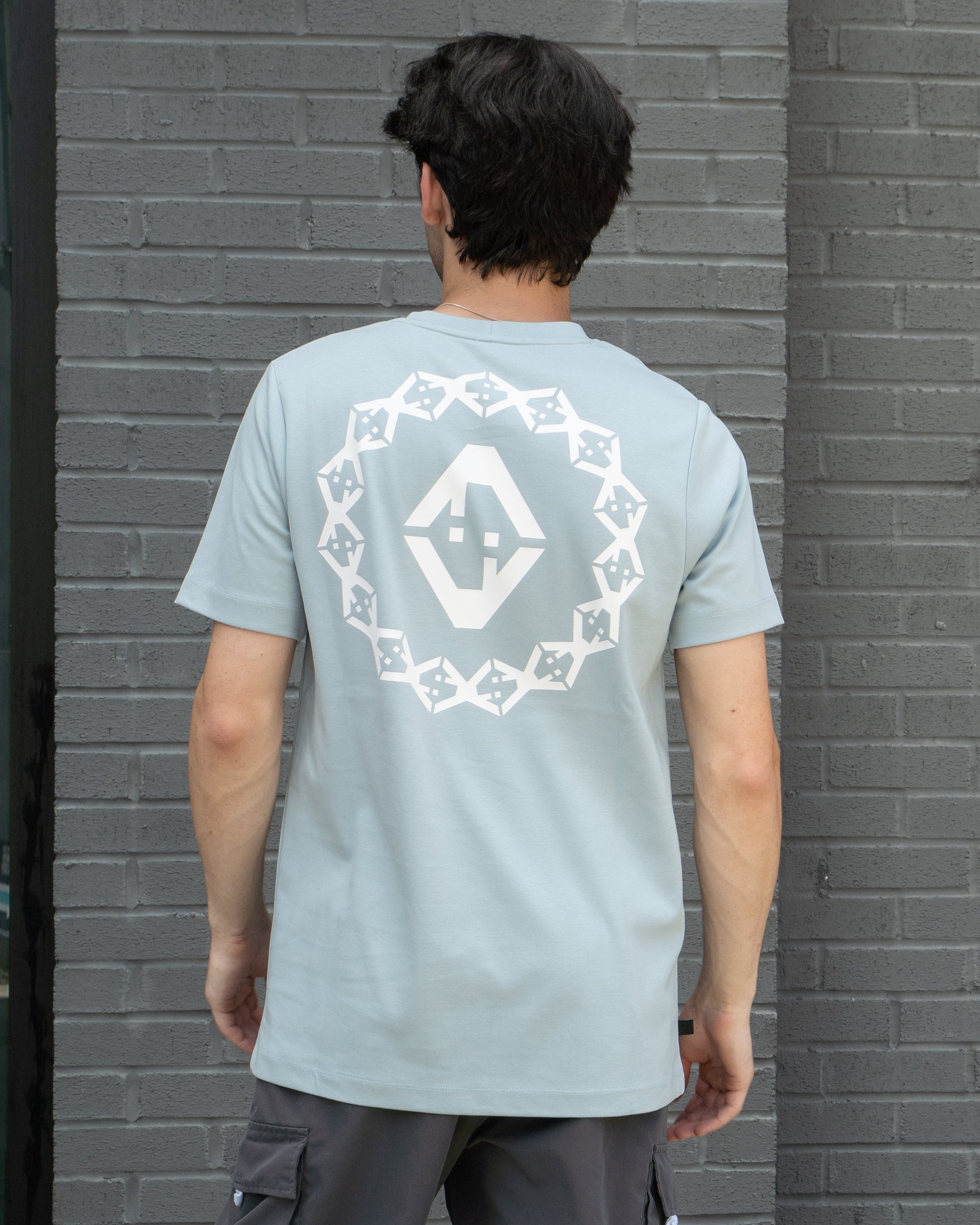 Person wearing a light blue t-shirt with a white graphic design on the back, standing against a gray brick wall.